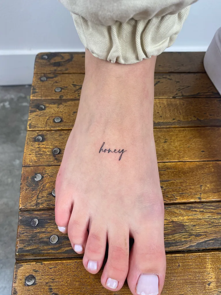 Honey script tattoo design for women, minimalist style, on the ankle, handwritten honey script across the top of the foot