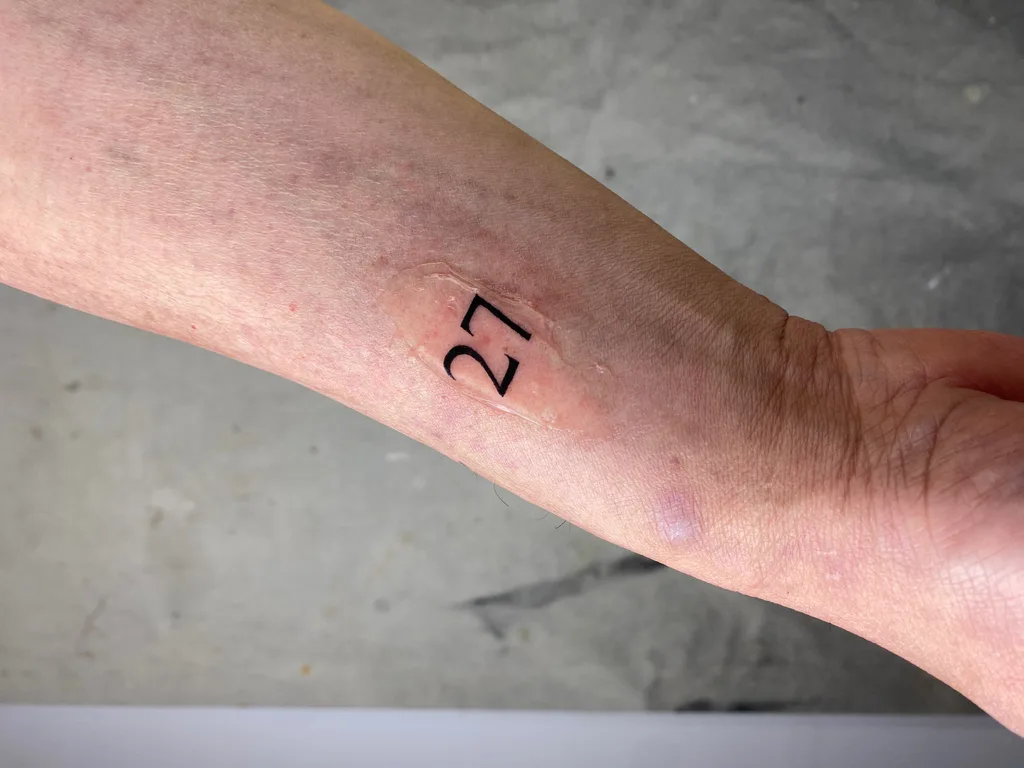 Number 27 tattoo design for women, minimalist style, on the forearm, small serif number 27 on the inner forearm